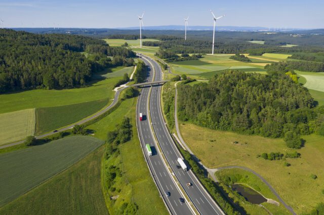 Autoroute à plusieurs voies avec camions en zone rurale, éoliennes en arrière-plan, vue aérienne.
éco-conduite